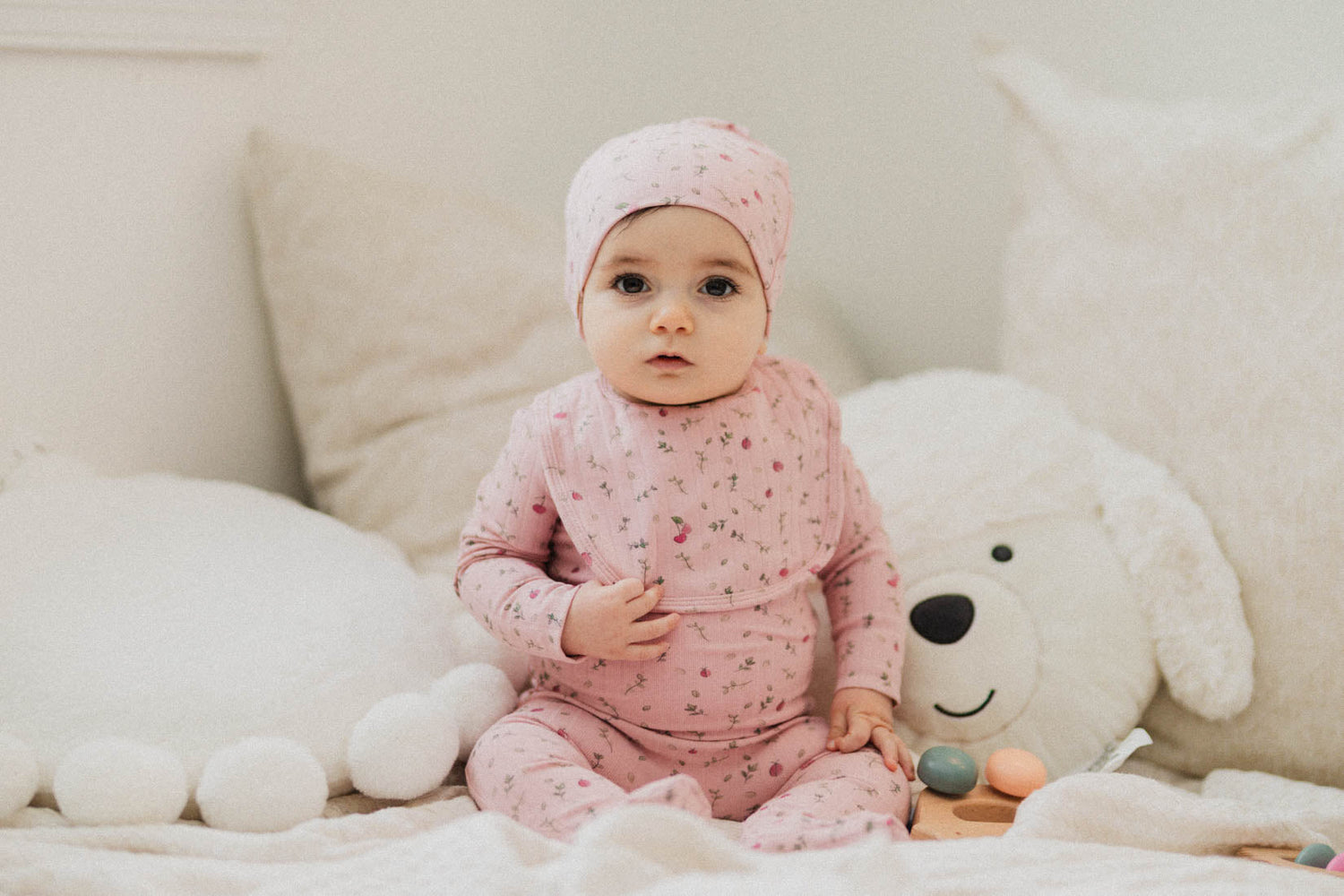 Adorable baby in a pink outfit with a matching hat, sitting on soft pillows with a toy near her.