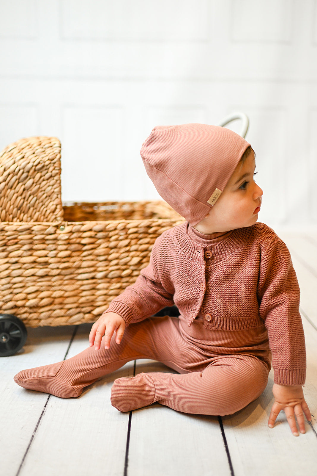 Baby girl in pink ribbed footie with mock neck and matching beanie, posing on a white floor.