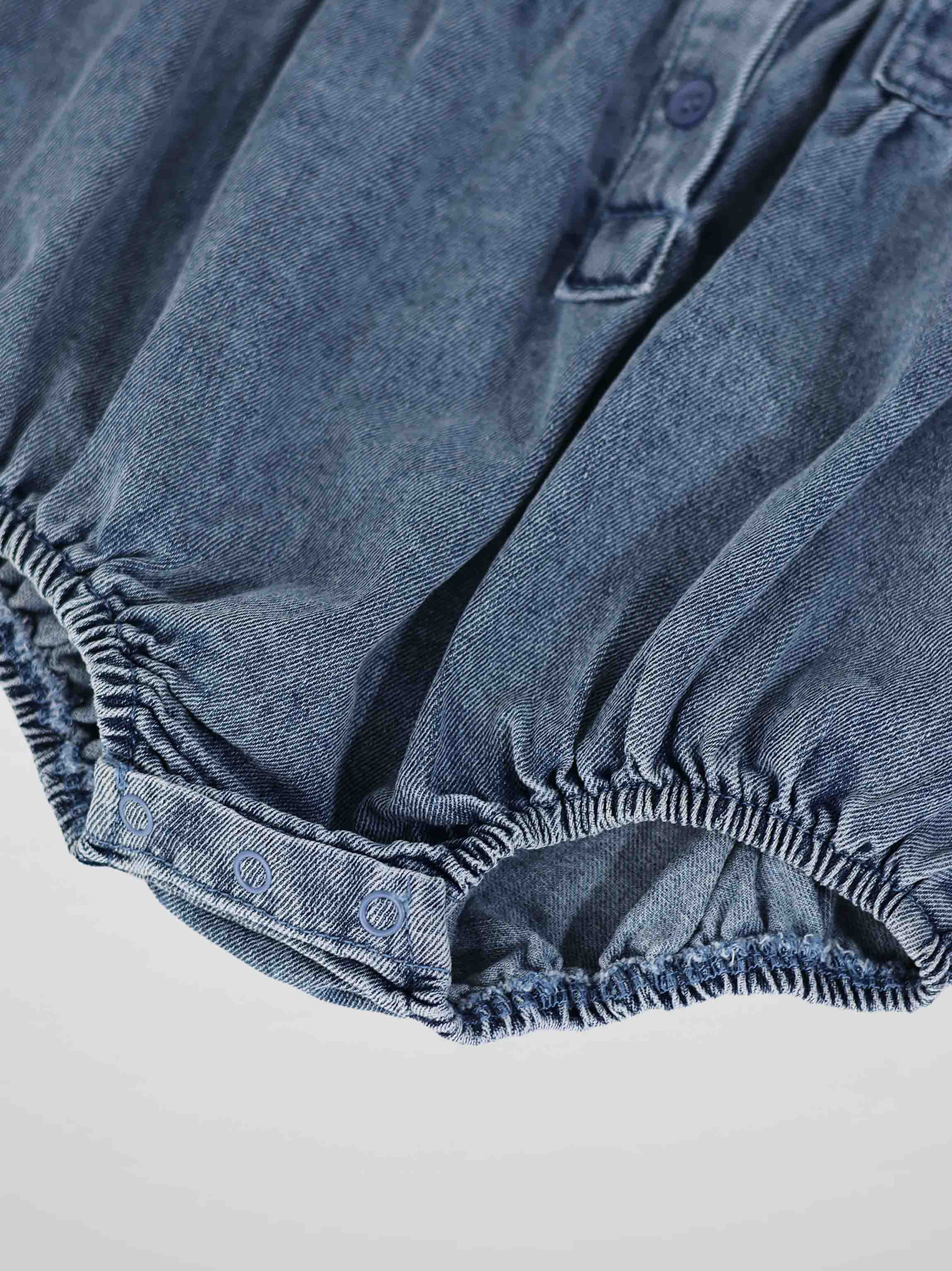 Blue denim romper leg opening with elastic gathers and metal snap closures.