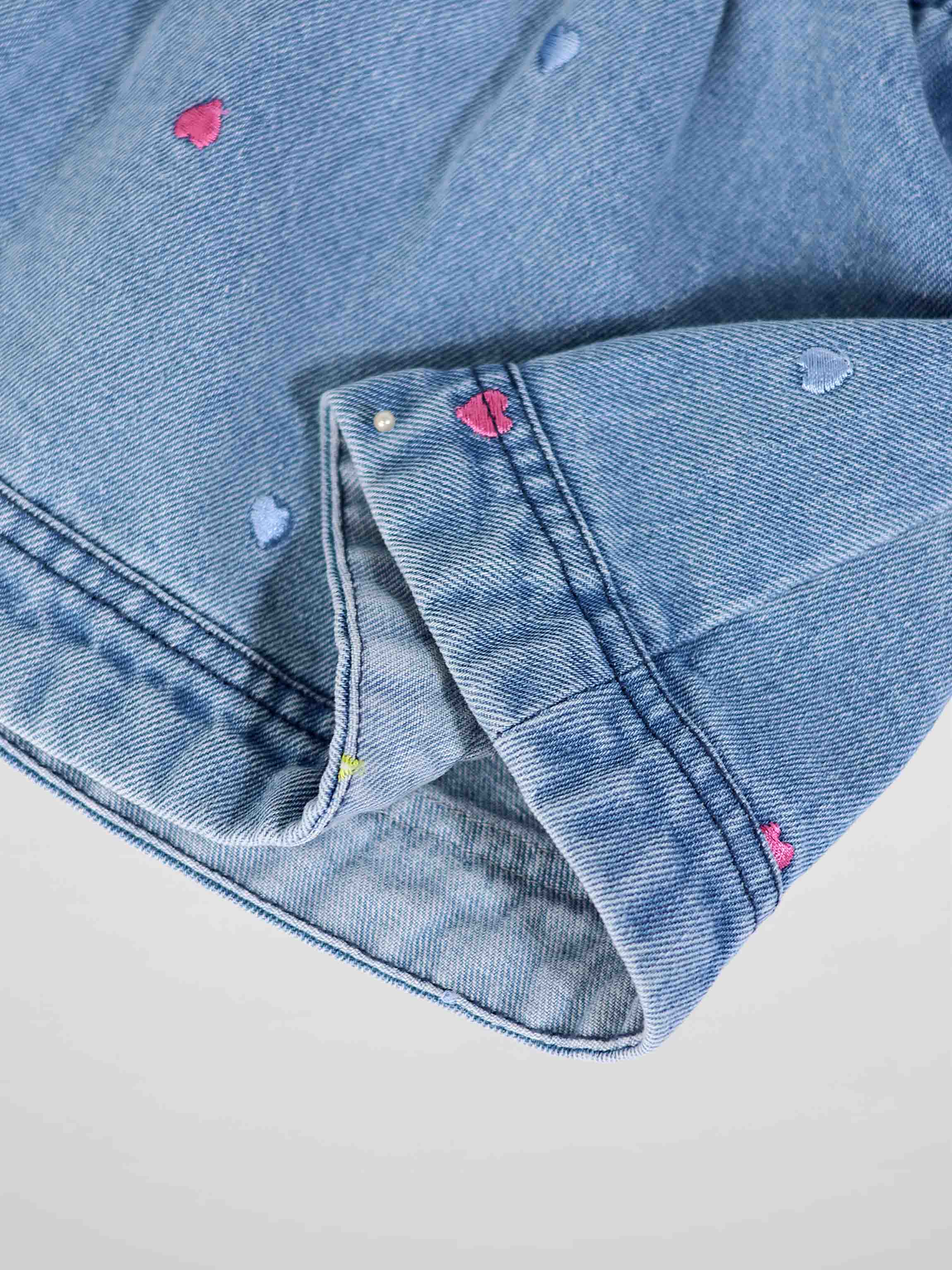 Close-up of light-wash denim with pink and blue heart embroidery on the fabric.