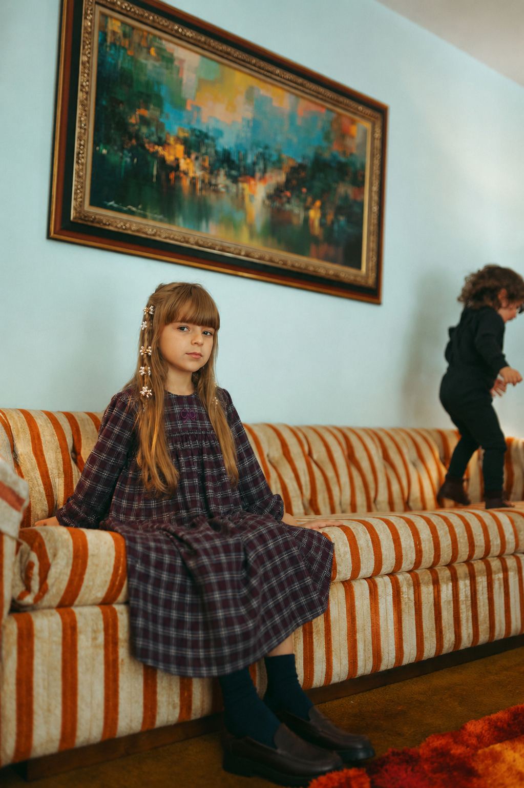 Plaid burgundy dress for girls, dressy style, worn by a seated child on a striped sofa.