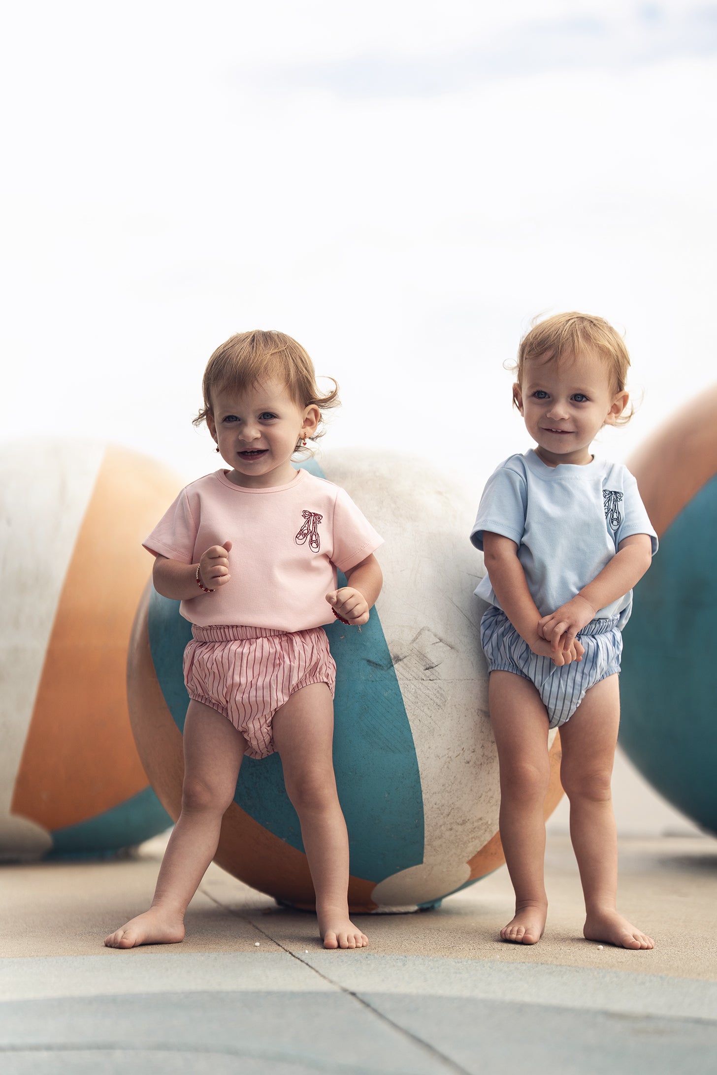 Two toddlers wearing striped bloomers; pink bloomers on the left, blue striped bloomers on the right, barefoot beside colorful balls.