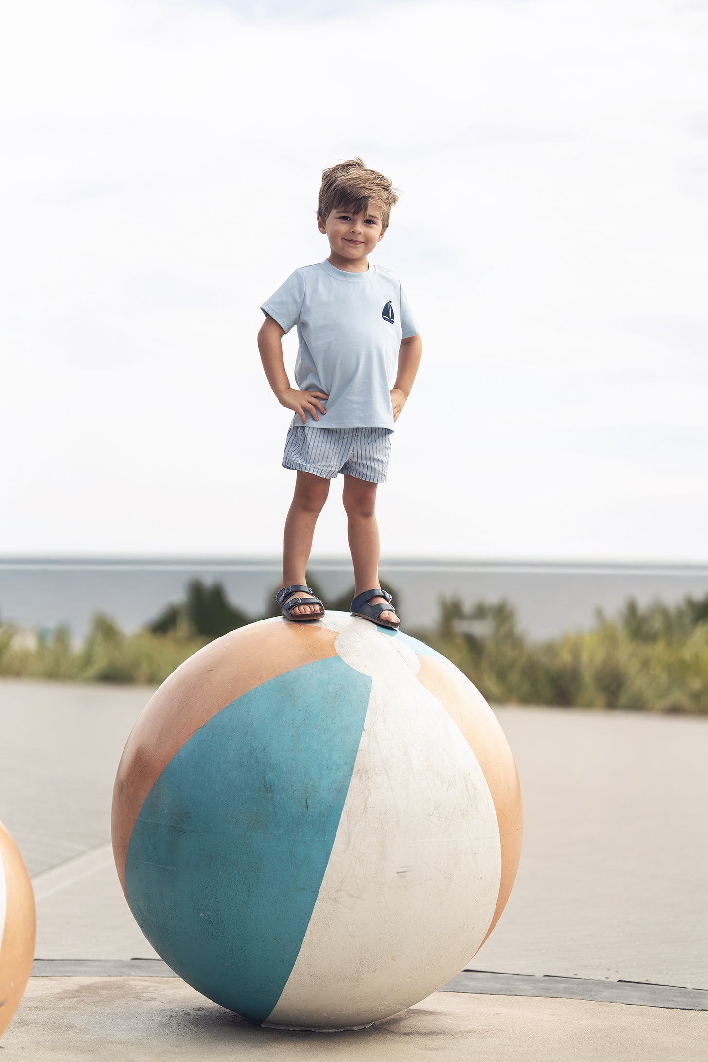 Young boy wearing light blue Short Sleeve Emblem Boxy T-Shirt stands on a large beach ball outdoors
