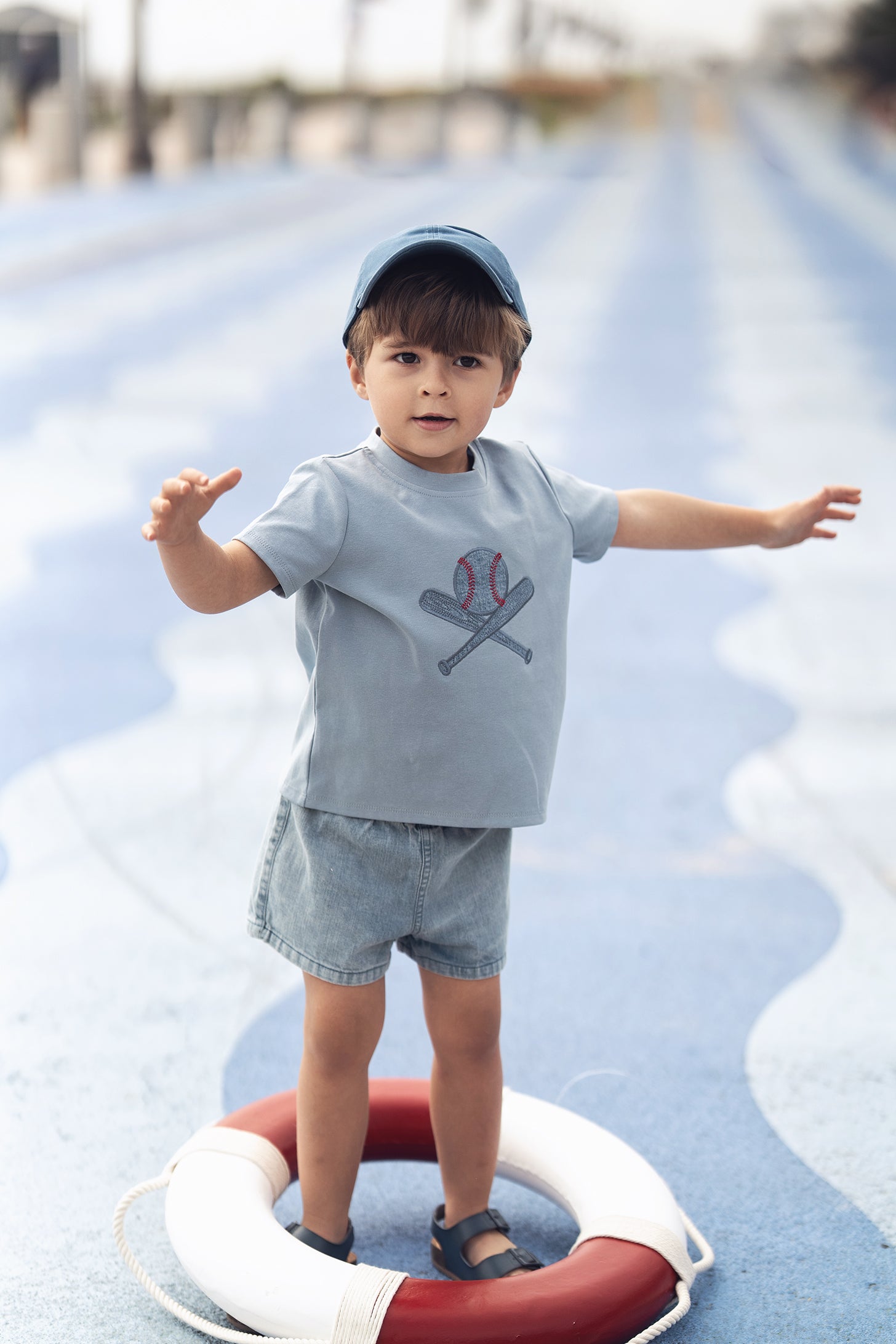 Child wearing a light blue Baseball T-shirt with crossed bats and ball logo, denim shorts, and blue cap.