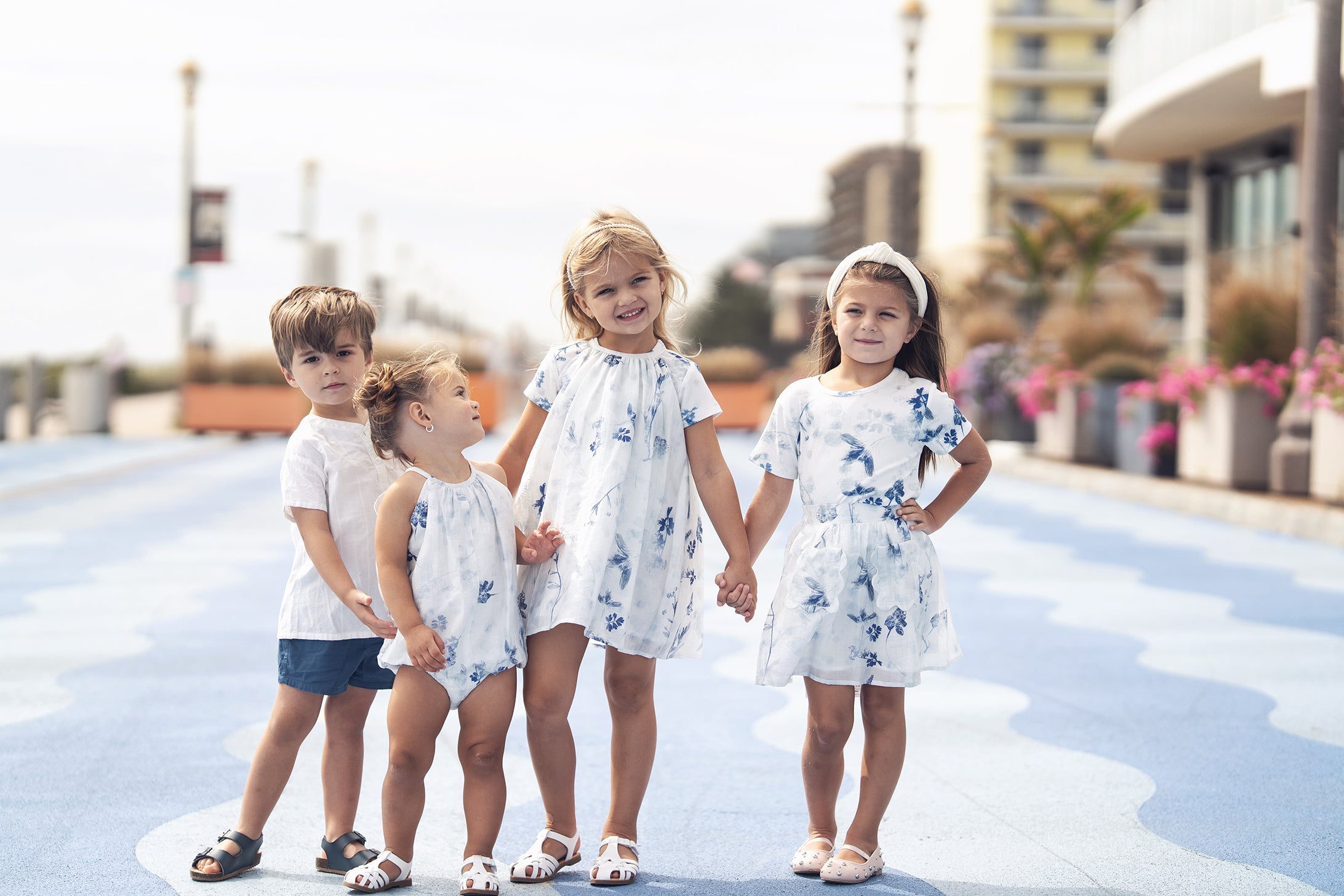 Children in white and blue floral outfits inspired by Blue Flower Skirt, walking hand in hand on a sunny boardwalk.