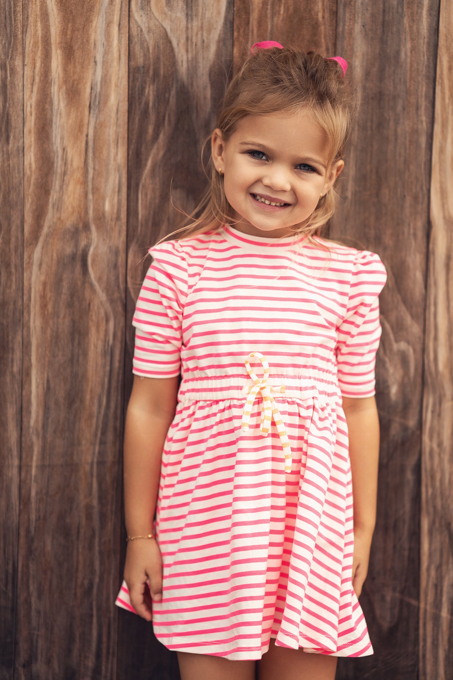 Smiling girl wearing neon pink and white striped dress with a yellow waist bow against a wooden backdrop.