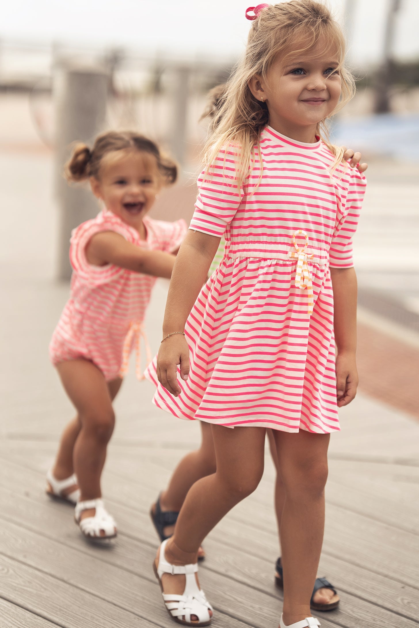 Two girls in neon pink striped dresses with 3/4 sleeves on a boardwalk.
