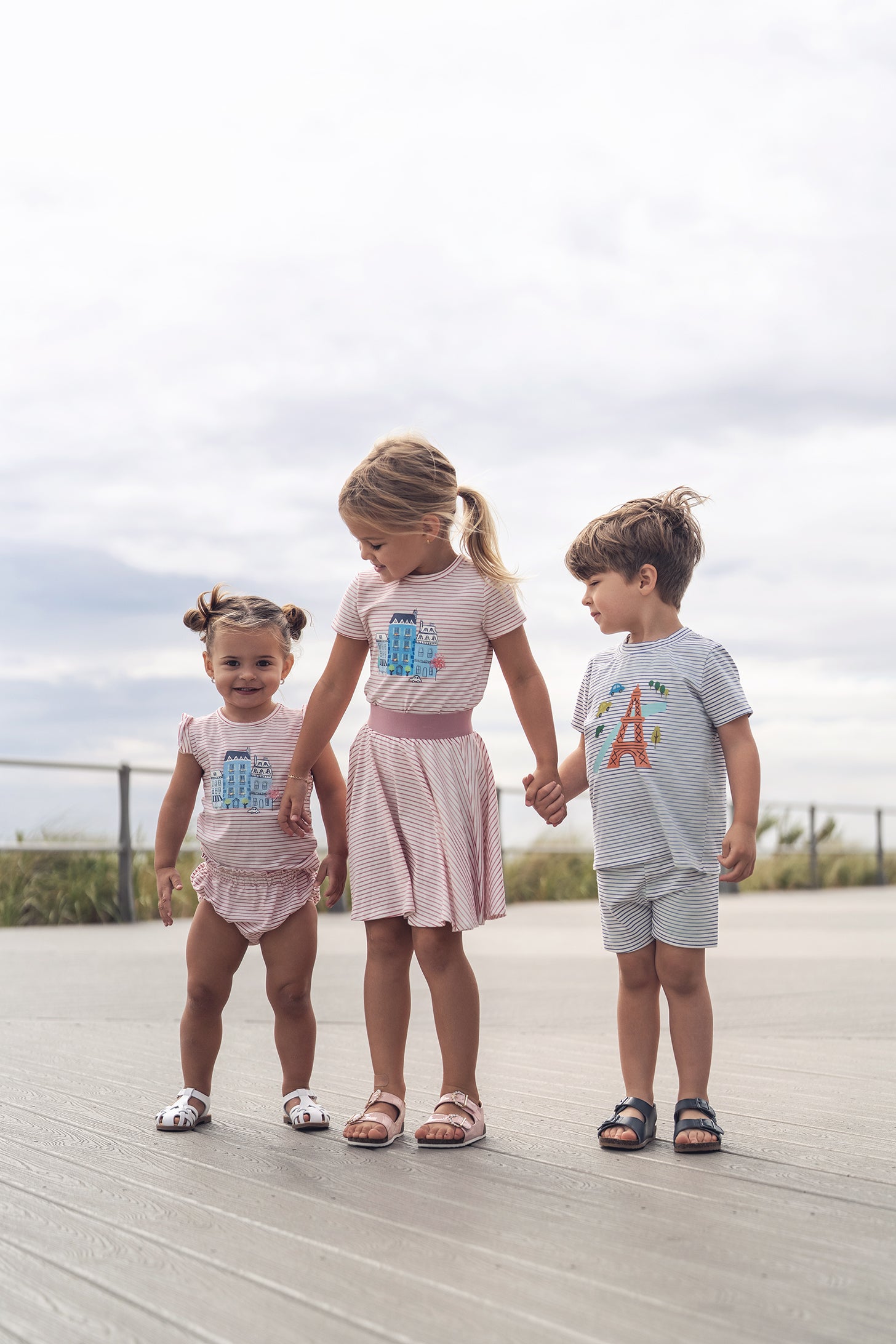 Three kids wear Eiffel Tower Shorts Set in pastel striped outfits on a boardwalk.