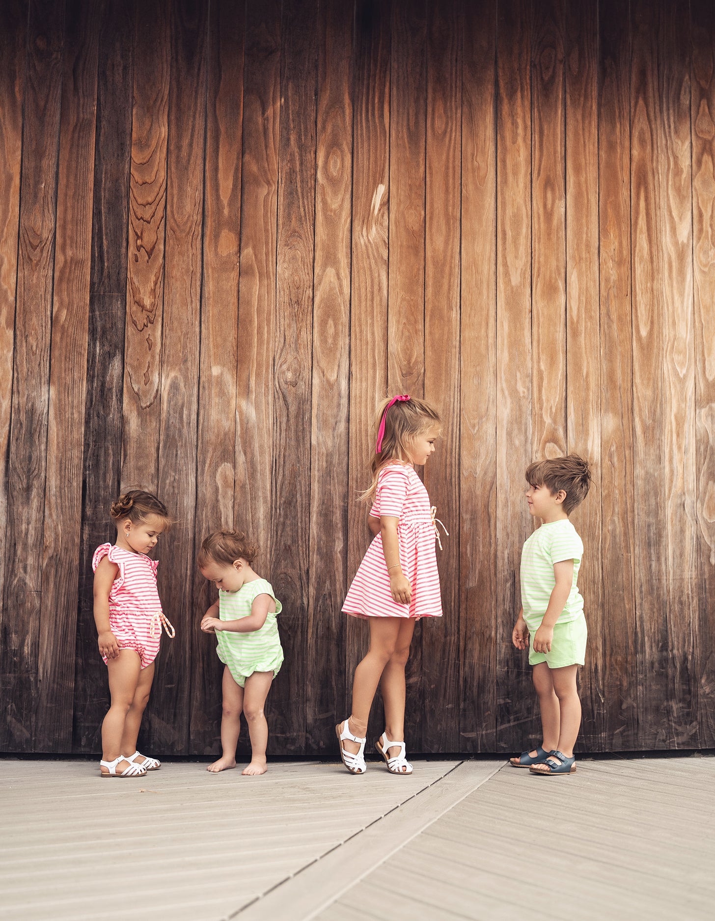 Kids in neon green striped romper and pink striped outfits pose against a weathered wooden wall.