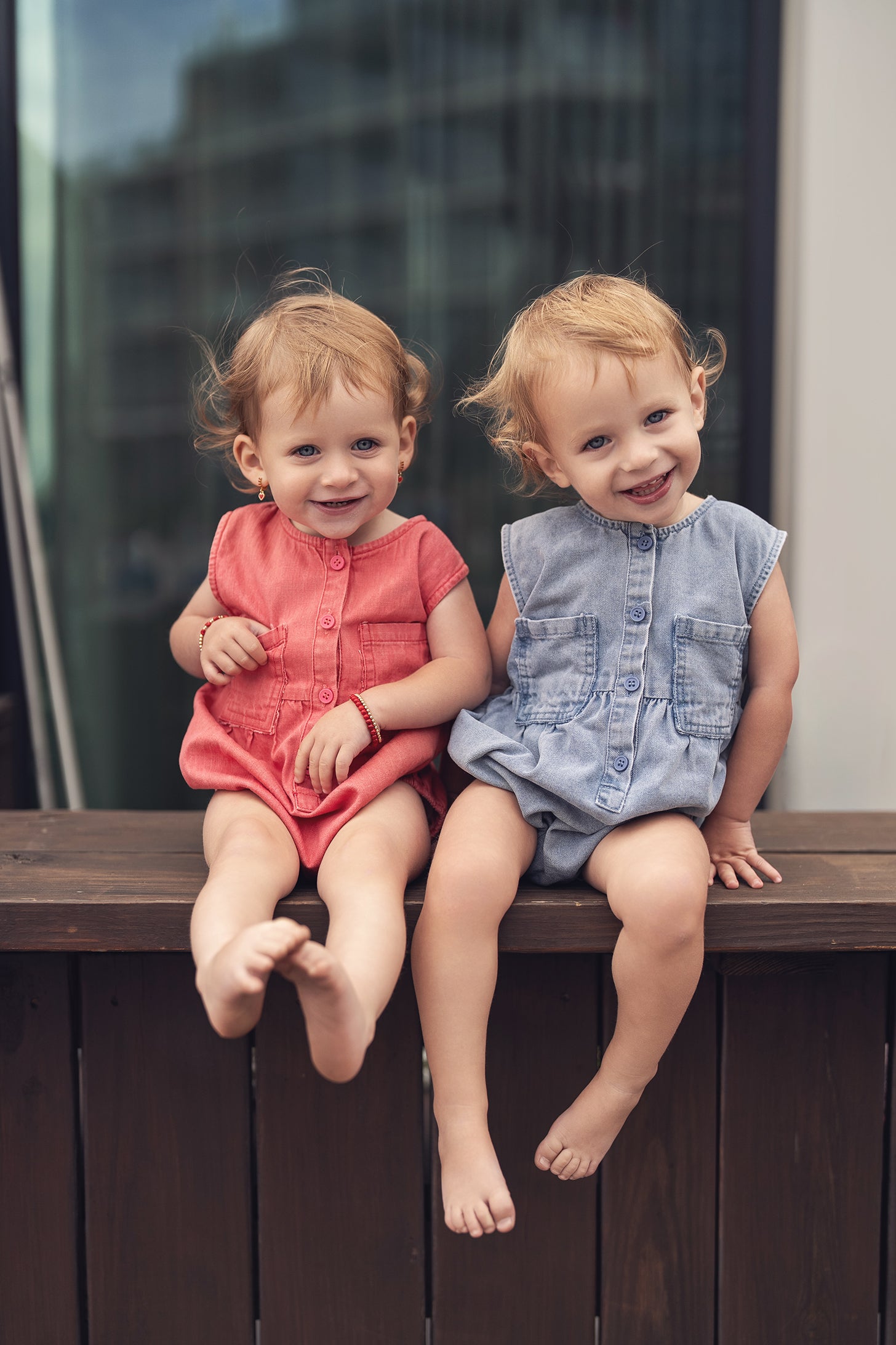 Two toddlers in a blue denim romper and a coral romper, smiling on a wooden railing.