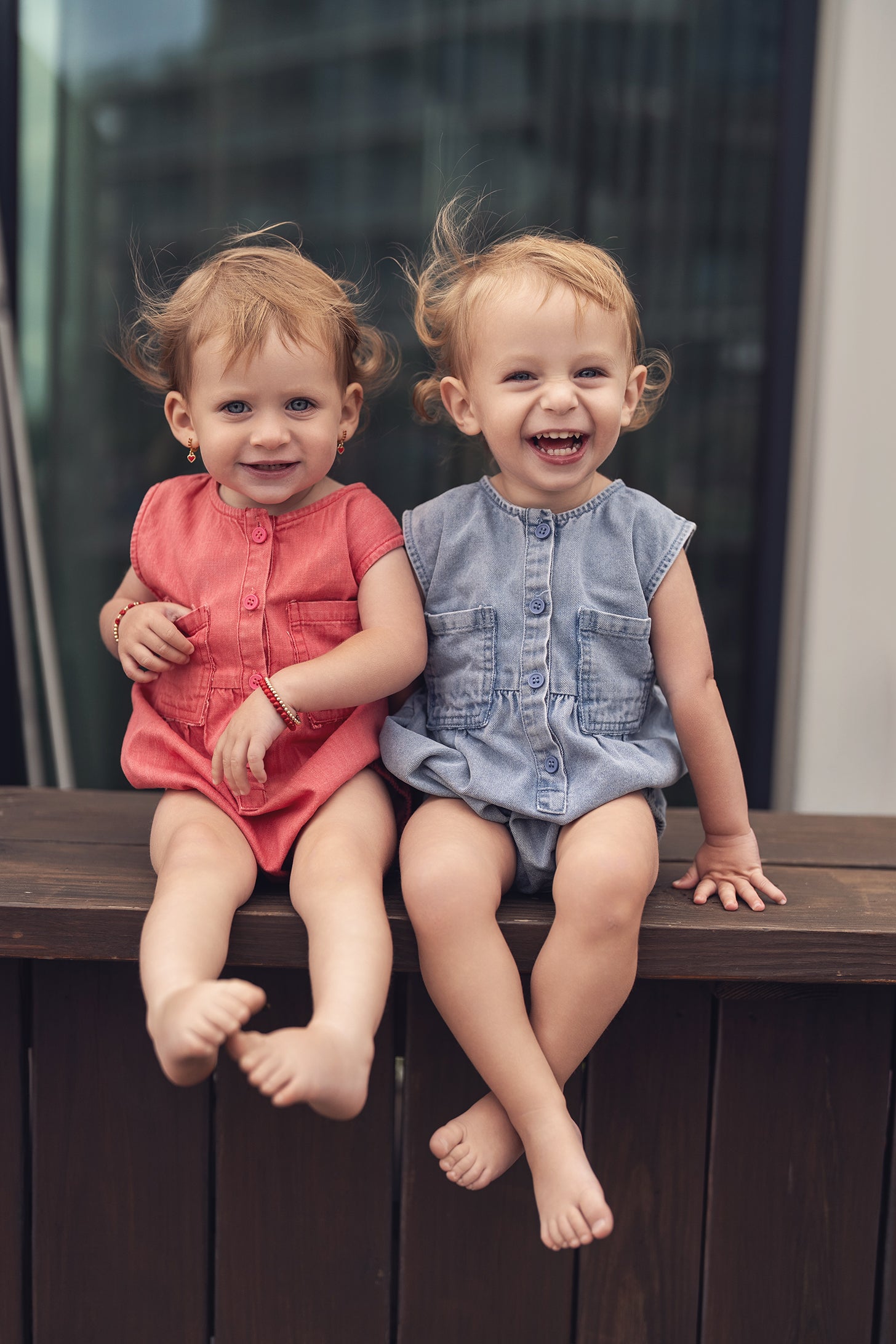 Two toddlers sit on a wooden railing in denim rompers; one in coral, one in blue denim, smiling.