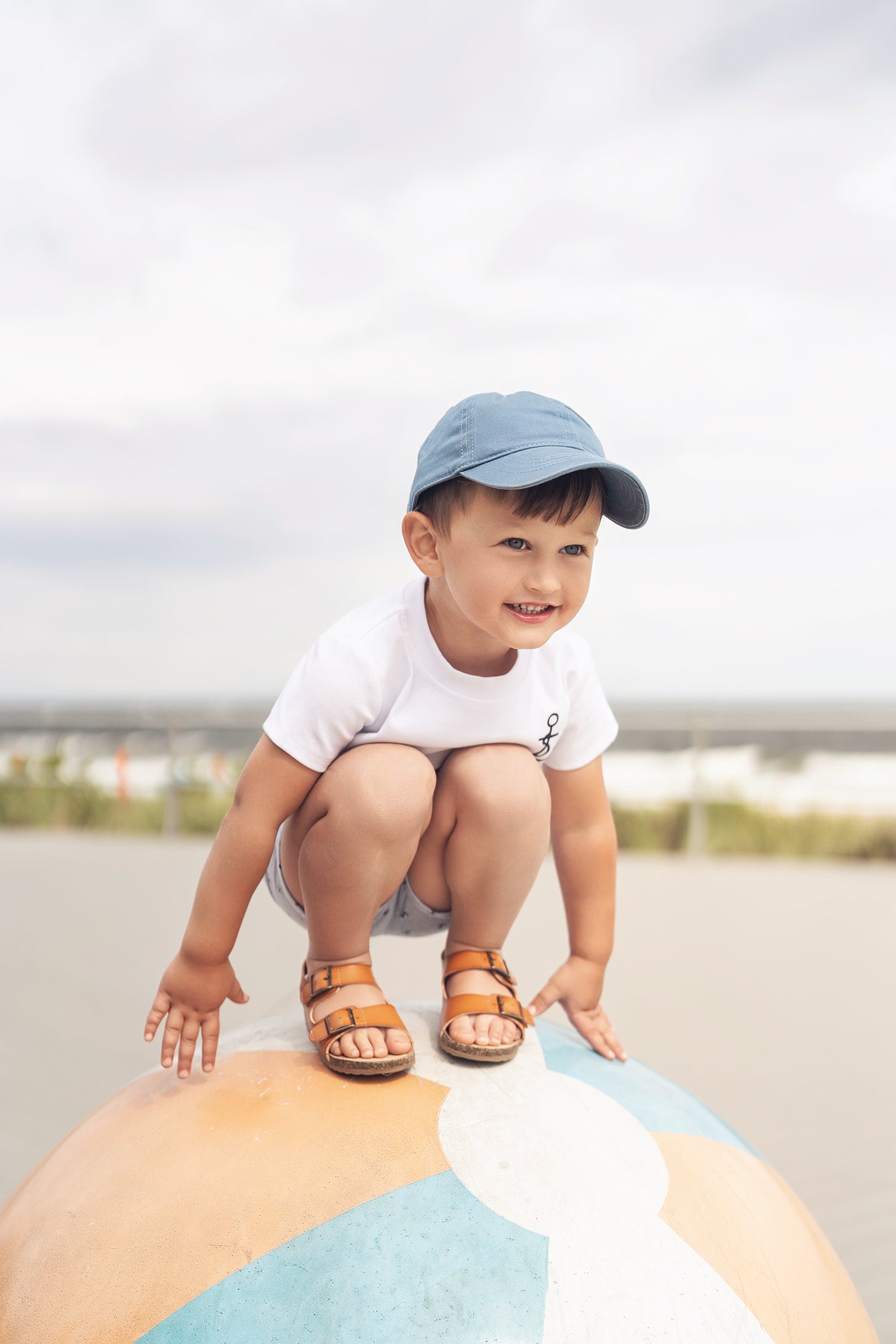 Young child wearing Embroidered Anchor Boxy Short Sleeve T-Shirt, squatting on a colorful beach ball at the shore.