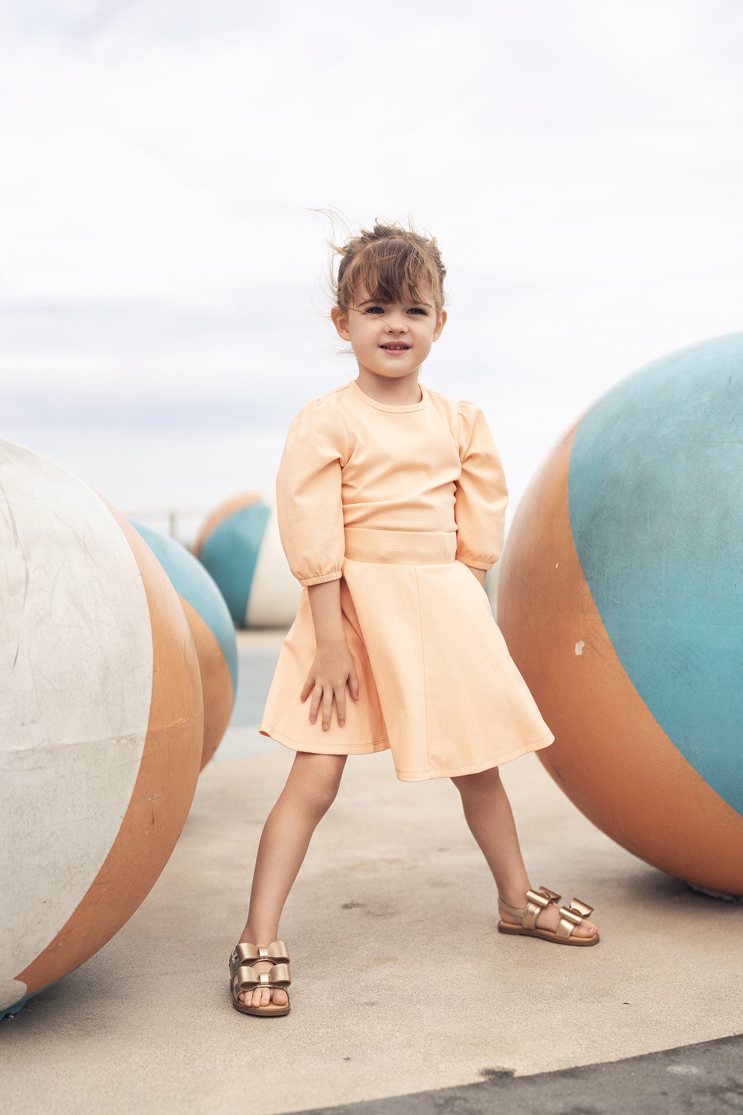 Peach 3/4 puff-sleeve dress on a young girl outdoors among oversized pastel spheres.