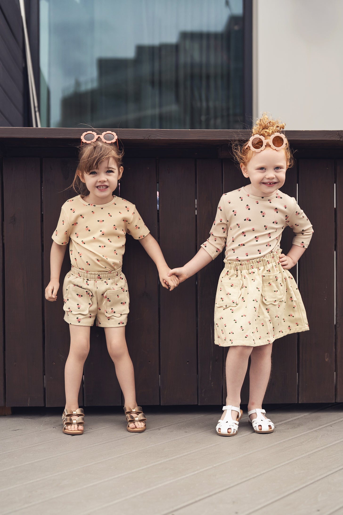 Two kids in tan floral outfits, one in shorts and the other in a matching floral skirt, holding hands with sunglasses.