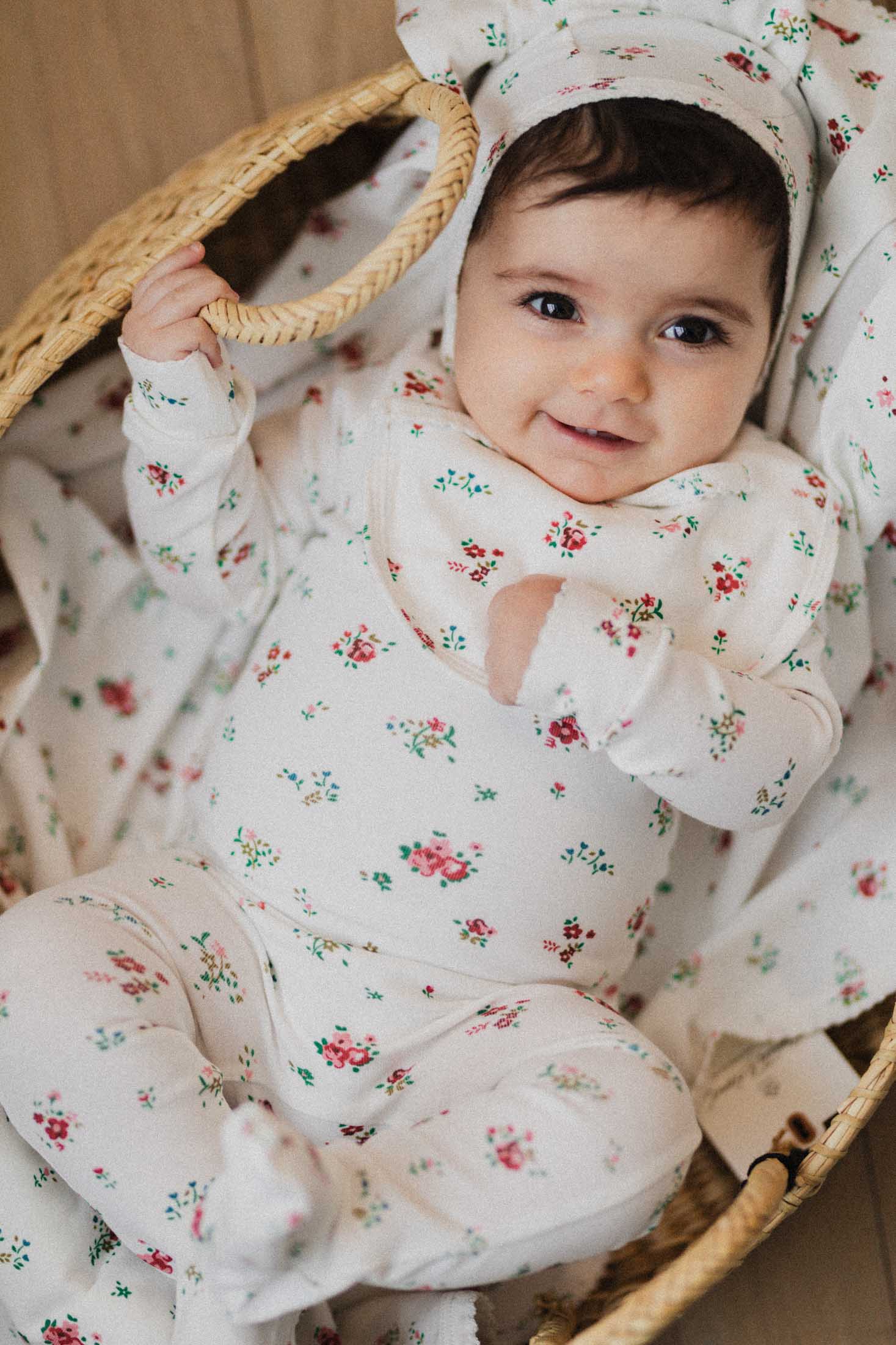 A smiling baby is lying in a woven basket, looking at the camera. They are wearing a white floral-patterned outfit and a matching hat.