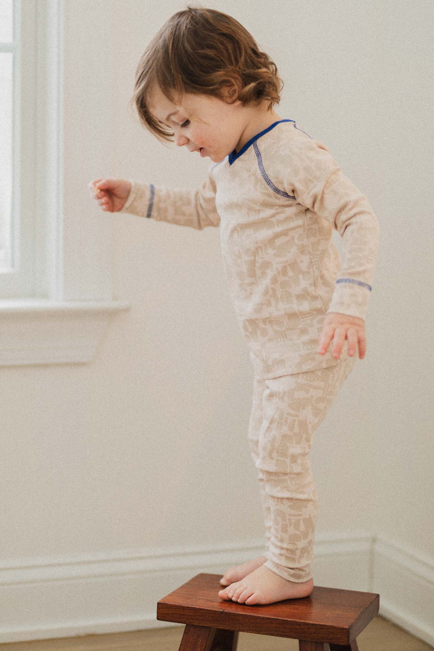 A young child stands on a small wooden stool, looking down. They are wearing light-colored pajamas with blue trim.