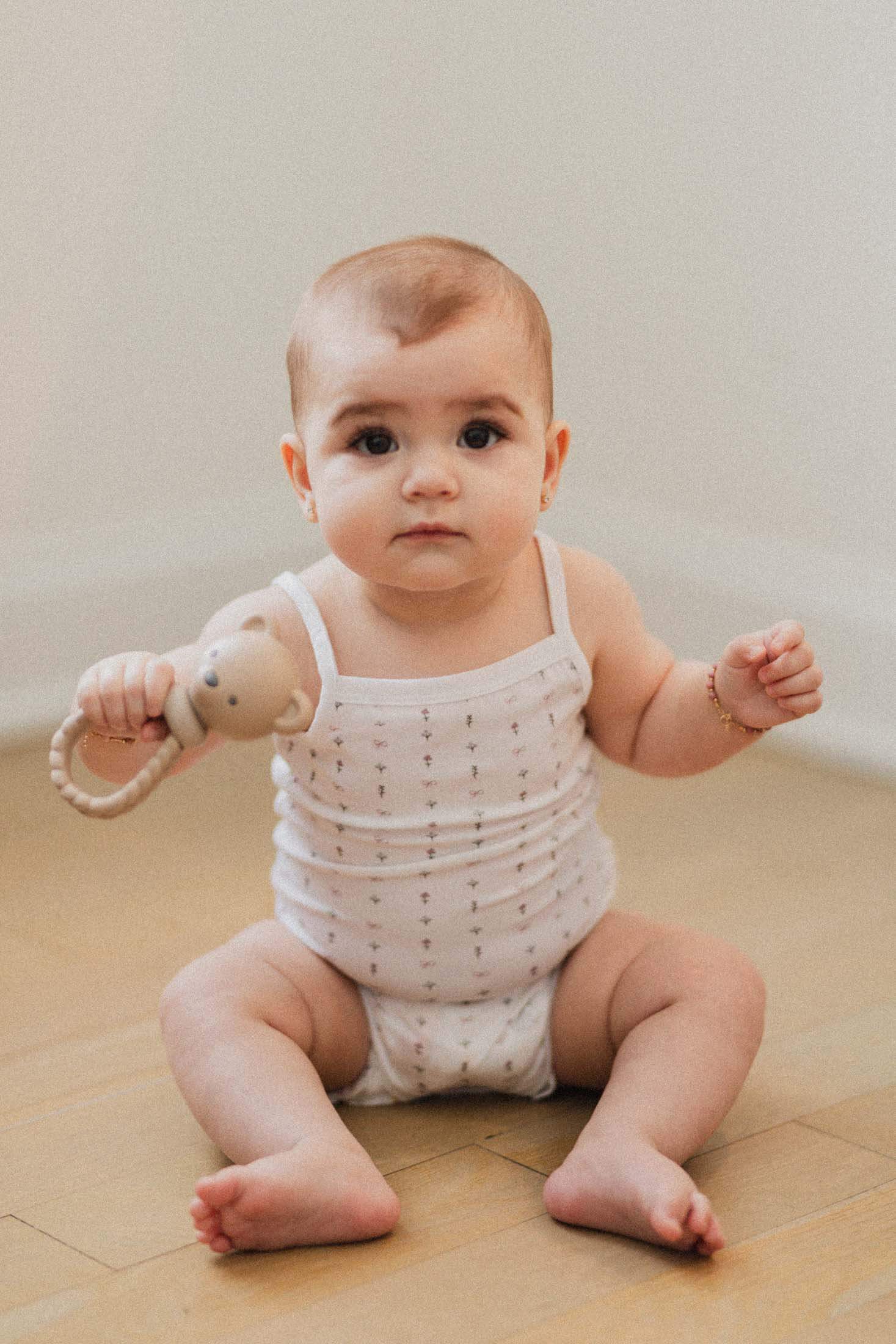 Adorable baby sitting on the floor in a white undershirt, holding a toy, showcasing soft infant clothing.