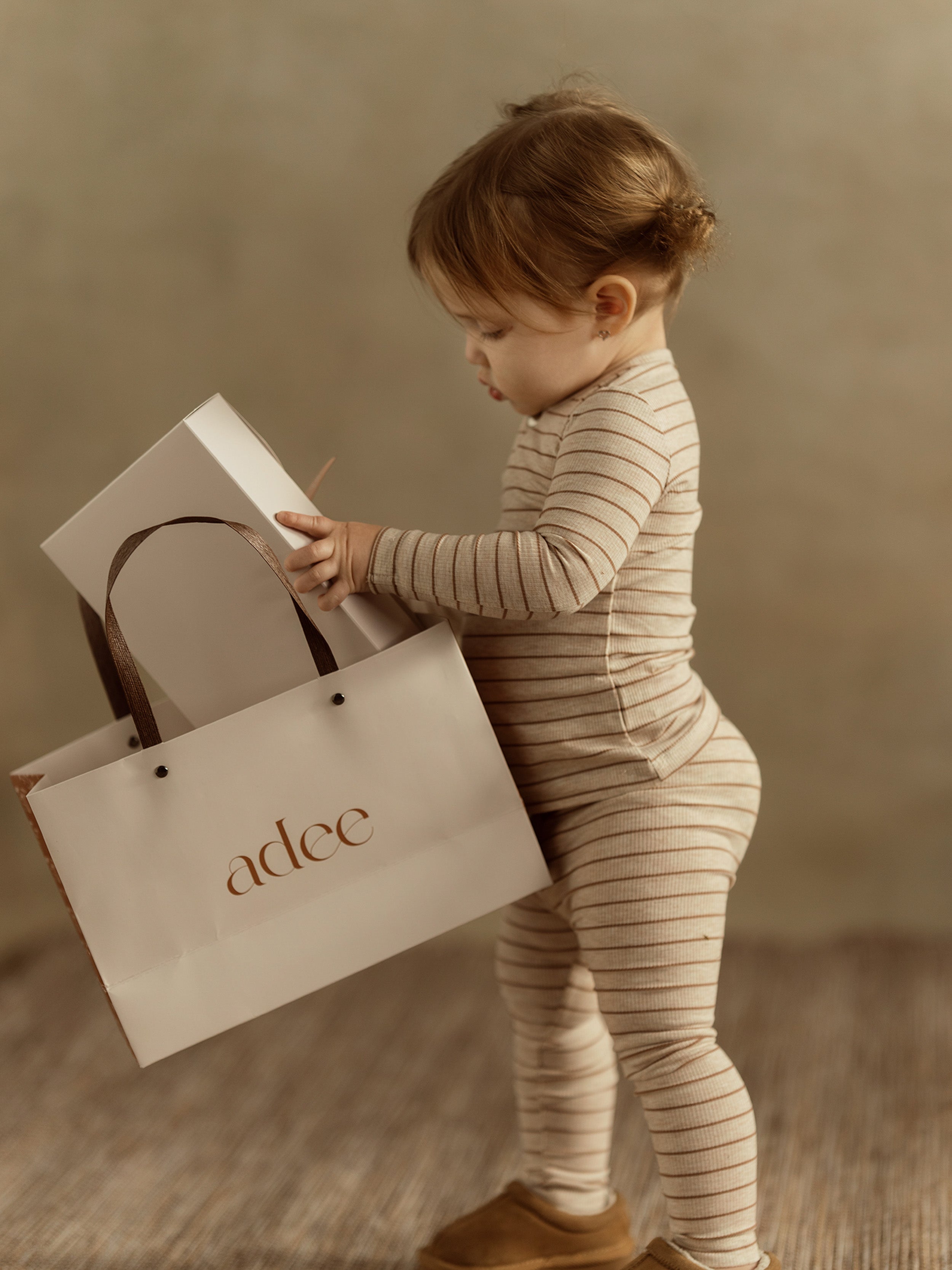 Toddler in striped outfit holding a gift bag, exuding joy in a warm, softly lit setting.