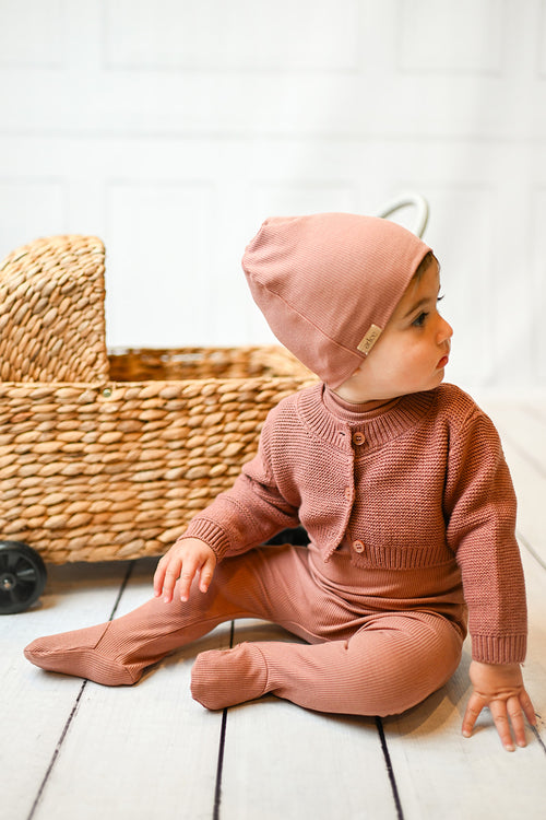 Baby girl in pink ribbed footie with mock neck and matching beanie, posing on a white floor.
