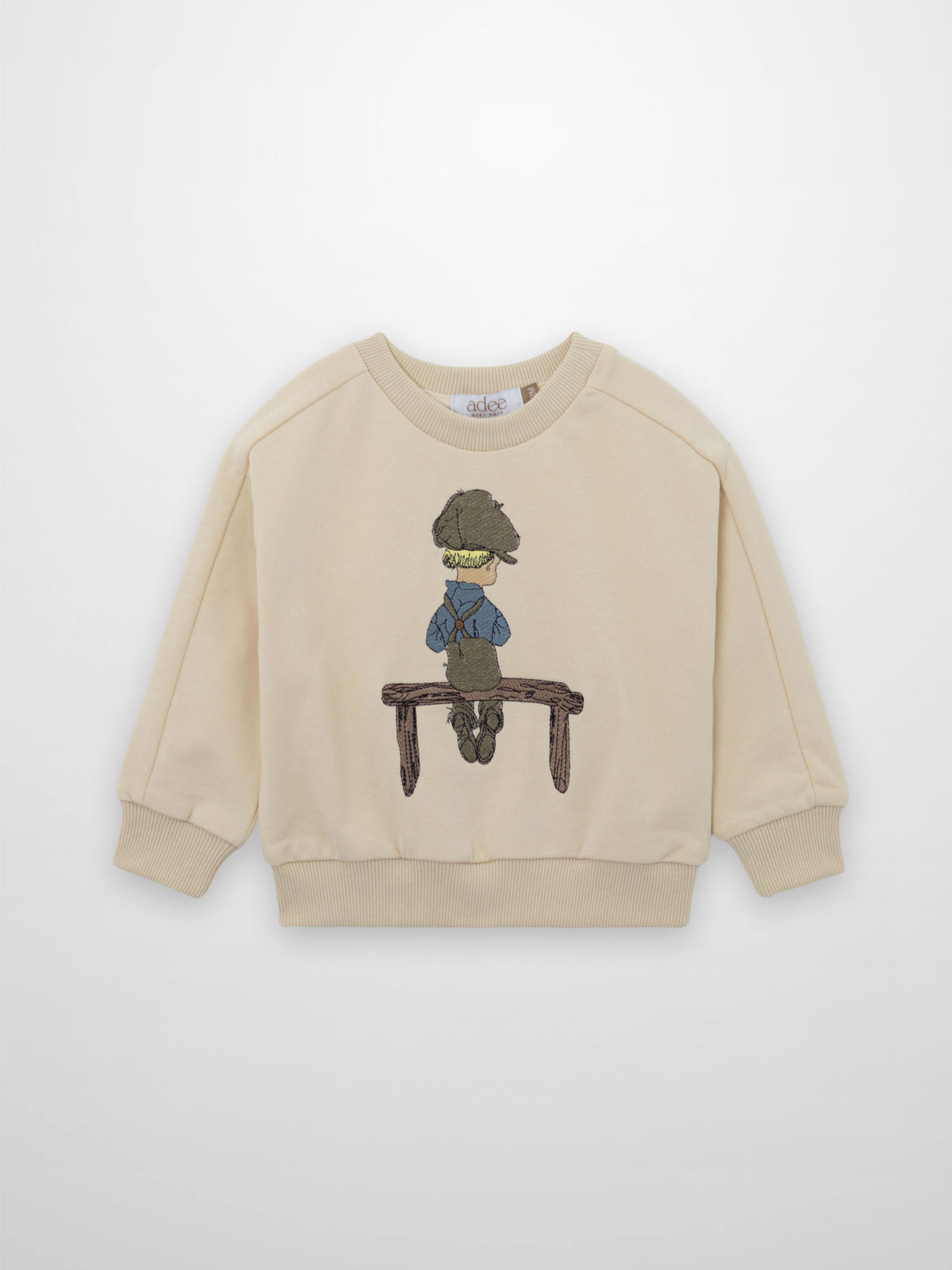 Beige embroidered boy sweatshirt shows a blond boy in a worn cap, blue shirt, brown overalls on a rustic bench.