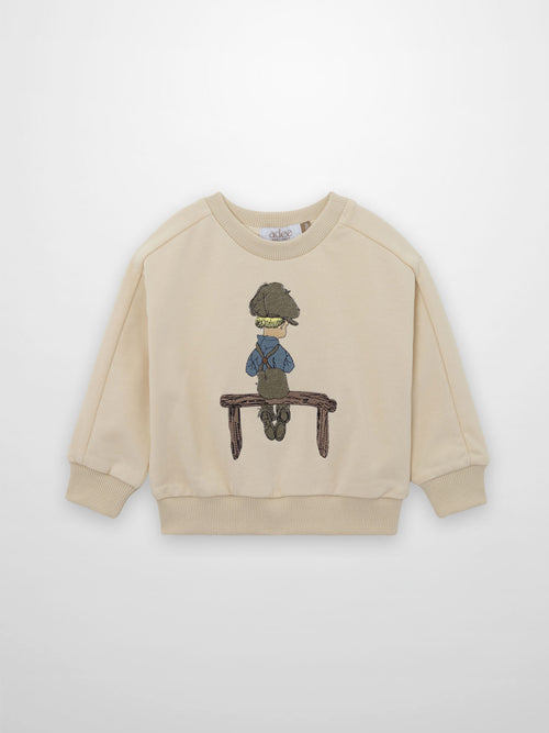 Beige embroidered boy sweatshirt shows a blond boy in a worn cap, blue shirt, brown overalls on a rustic bench.