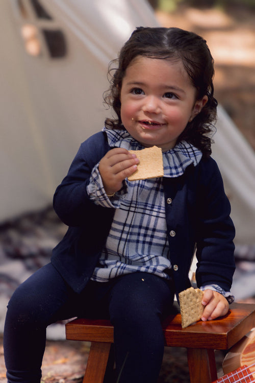 Plaid Shirt - Blue worn by a young girl in a navy cardigan, sitting on a bench and eating crackers.
