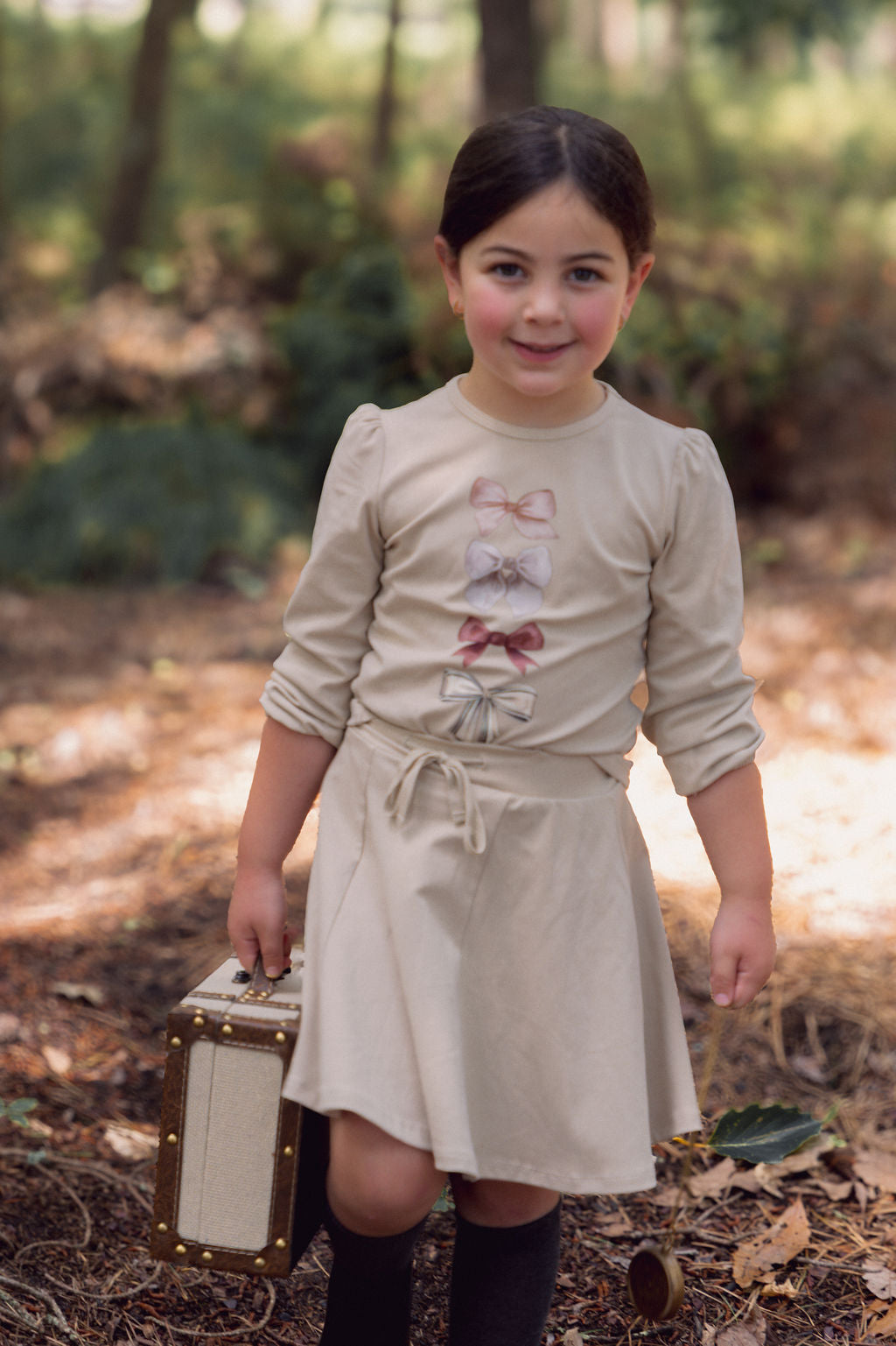 Young girl wearing a taupe six-panel skirt with a tied waist, outdoors in a forest, carrying a vintage suitcase.