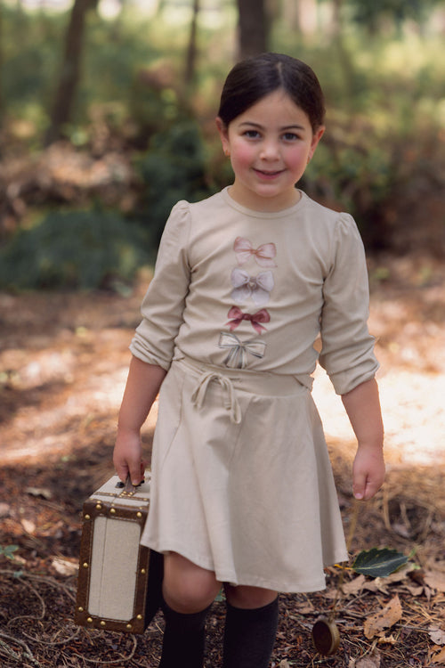 Young girl wearing a taupe six-panel skirt with a tied waist, outdoors in a forest, carrying a vintage suitcase.