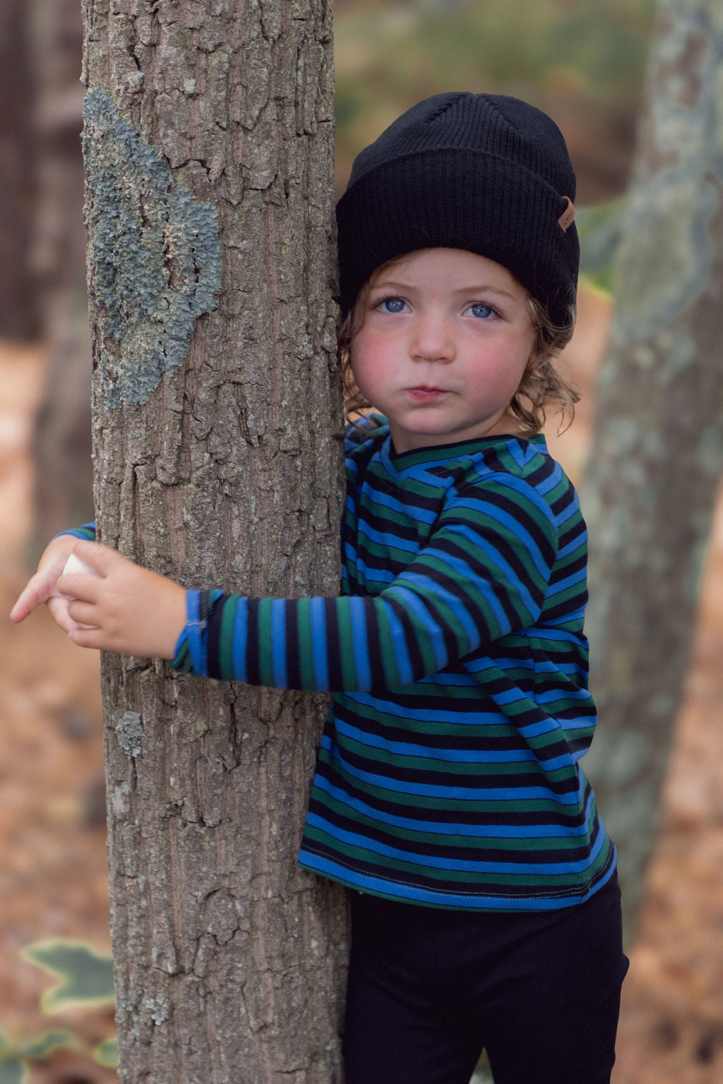 Child wearing blue striped long-sleeve crew-neck T-shirt with green and black stripes, hugging a tree outdoors.