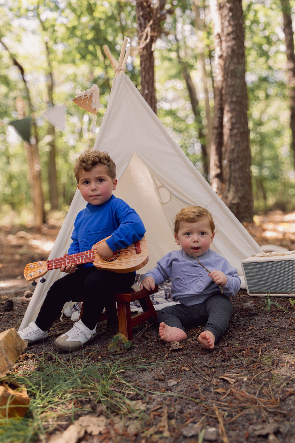 Two kids outdoors in a forest wearing a grey Bike Sweatshirt (minimalist, warm) with a toy guitar and a teepee tent