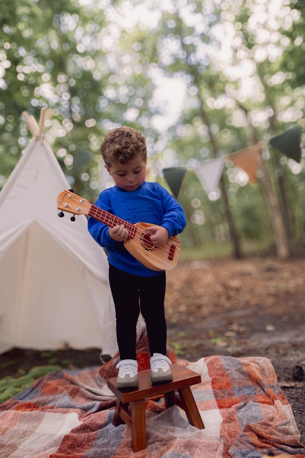 Toddler in royal blue Bike Sweatshirt stands on a stool, playing a small wooden guitar outdoors.