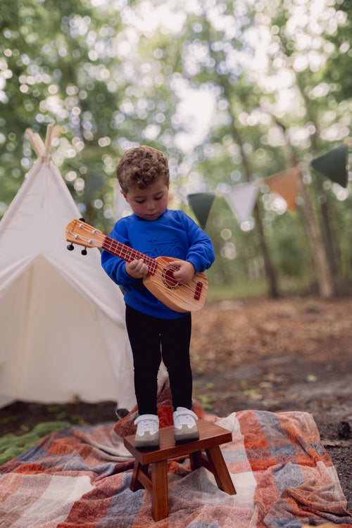 Toddler in royal blue Bike Sweatshirt stands on a stool, playing a small wooden guitar outdoors.