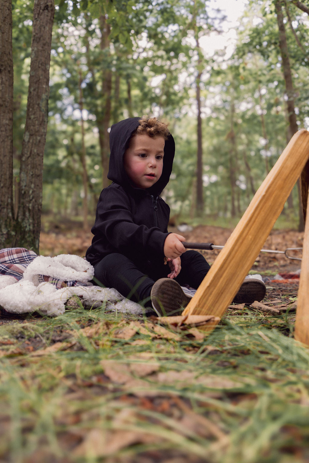 Young child in a black thin-rib zip hoodie sits on a blanket in a forest, playing with a wooden ramp.