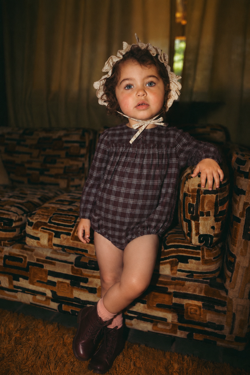 Plaid burgundy Adee girls romper dressy outfit worn by a toddler with beige lace bonnet and burgundy boots.