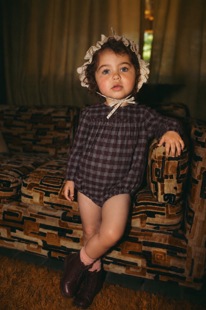 Plaid burgundy Adee girls romper dressy outfit worn by a toddler with beige lace bonnet and burgundy boots.