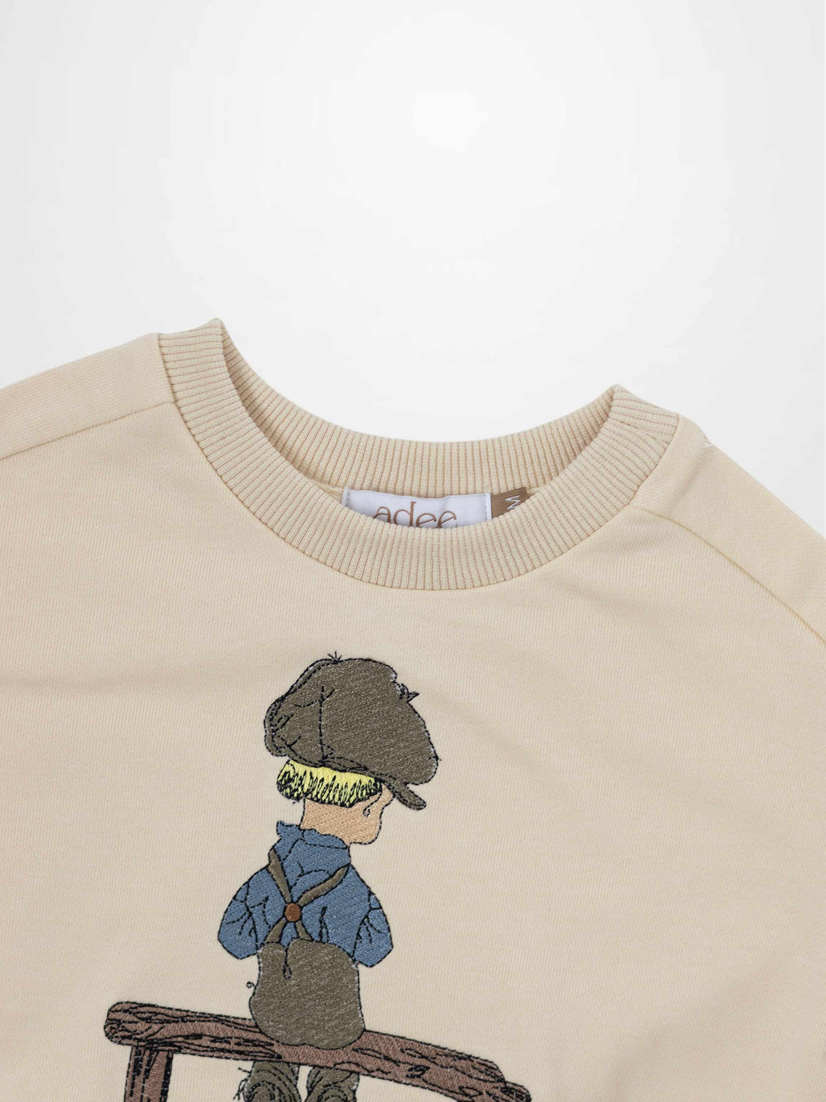 Beige embroidered sweatshirt with a blond boy in a cap, blue shirt and brown overalls sitting on a wooden bench.