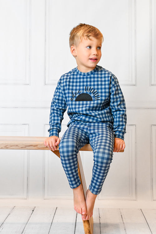 Boy wearing blue gingham pajamas with sun graphic, sitting barefoot on a wooden bench.