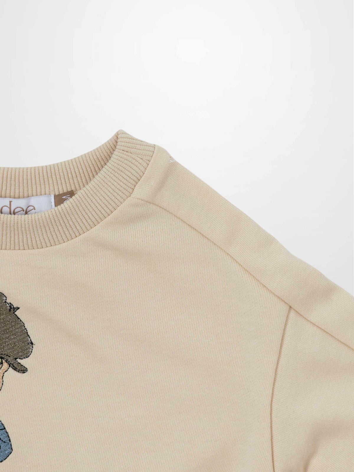 Beige embroidered boy sweatshirt with a blond boy in a worn cap, blue shirt, brown overalls on a rustic wooden bench.