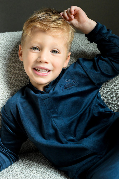 Smiling boy in navy velour bomber top, cozy on a gray sofa.