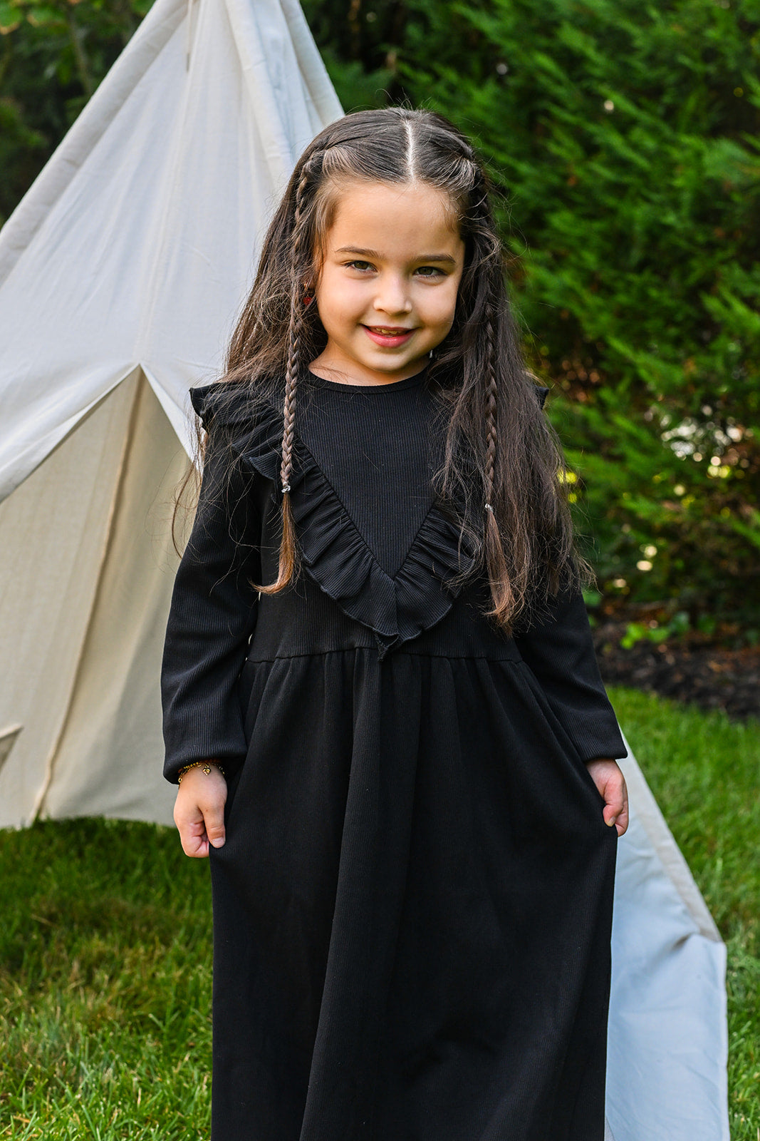 Girl in a black ribbed dress with a V-front ruffle, standing outdoors beside a tent.