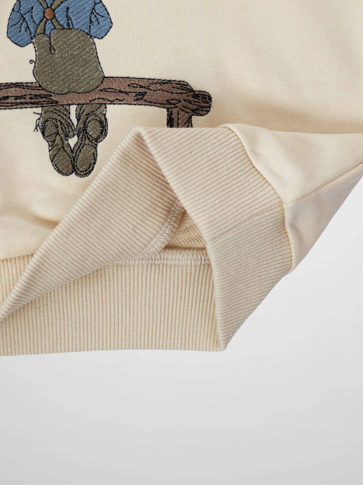Beige sweatshirt with embroidered blond boy in a worn cap, blue shirt and brown overalls on a rustic wooden bench.
