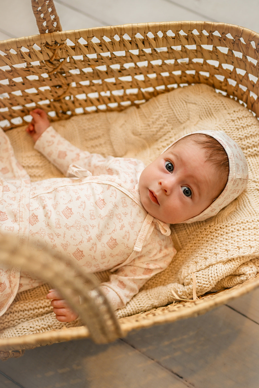 Little Storybook Stretchy - Pink baby jumpsuit worn by a baby girl resting in a woven basket on a knitted blanket.