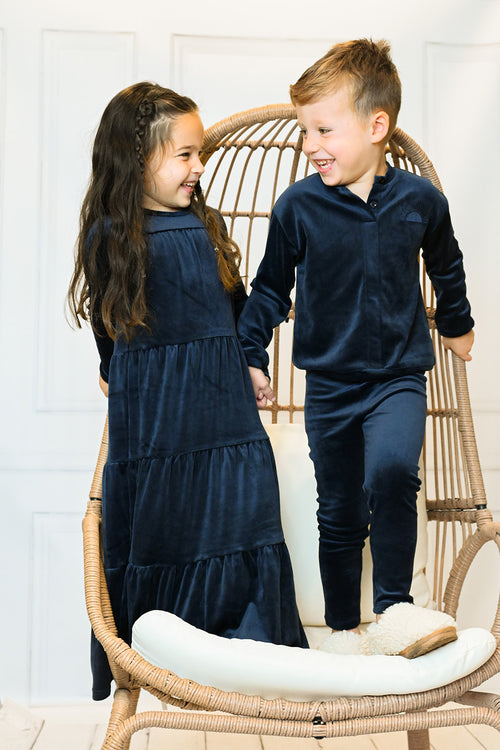 Two children in navy velour outfits, girl in tiered dress and boy in matching velour set, smiling indoors.