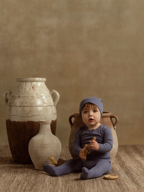 Baby wearing blue wide ribbed beanie, sitting on a woven rug beside ceramic pots.