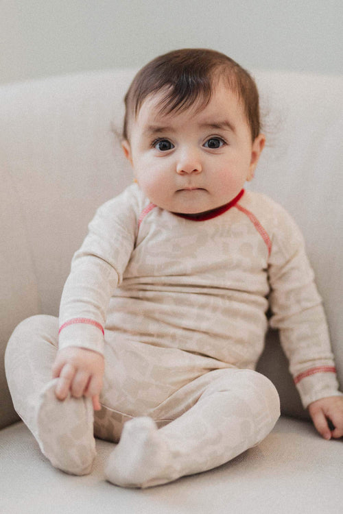 Cream abstract-print stretchy footed jumpsuit with red trim and V-neck, baby seated on sofa.