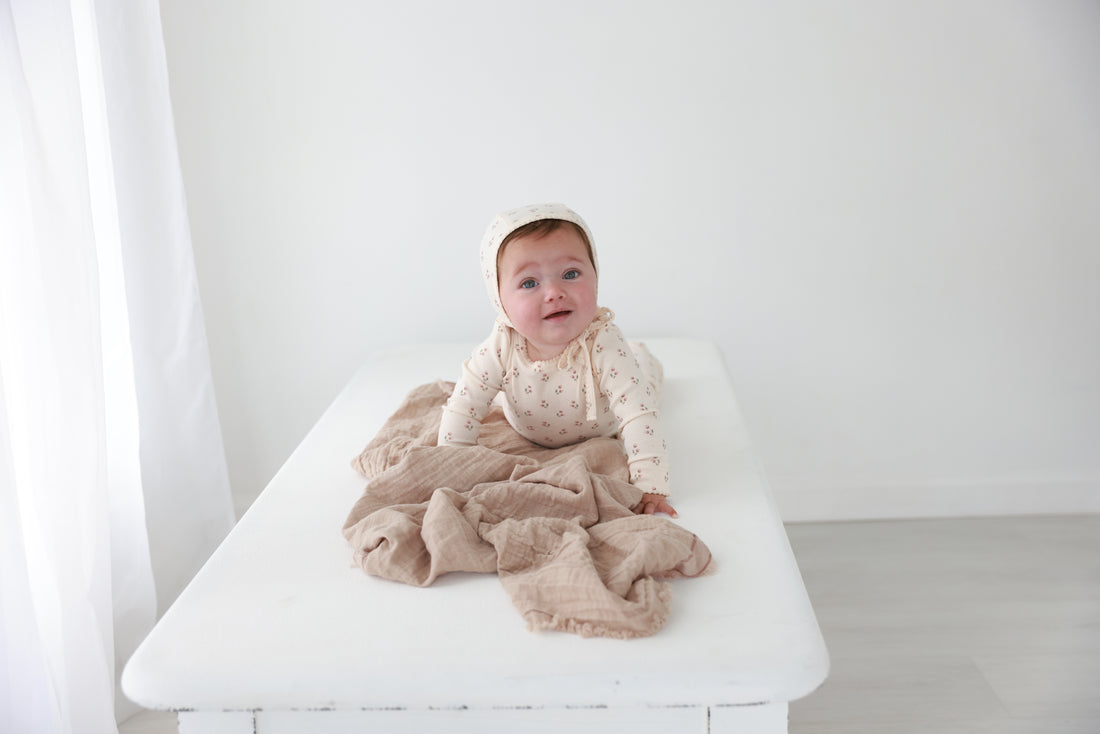 Tiny Tulip layette set for girls by Adee, baby in cream floral bonnet and matching bodysuit on a white bed.