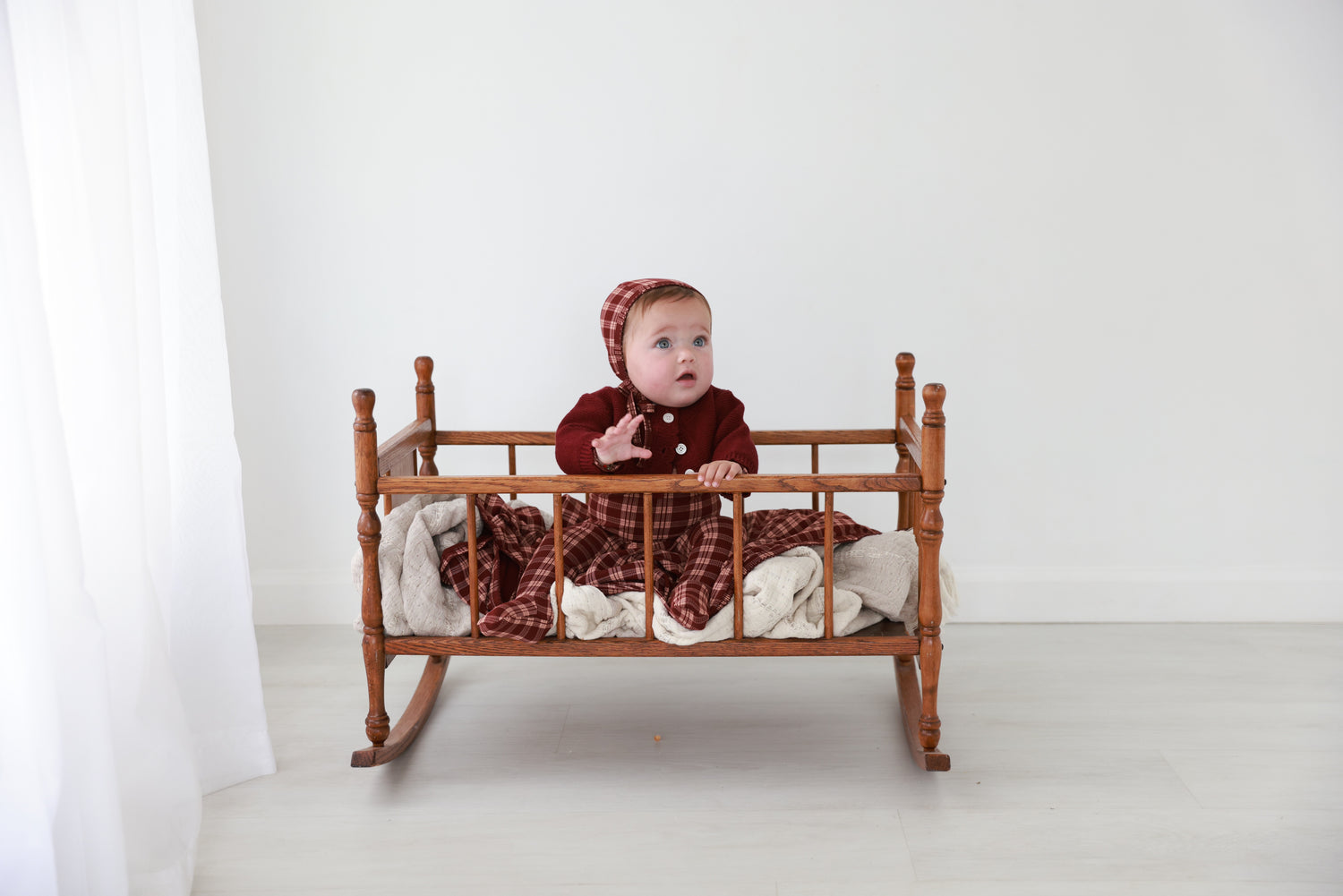 Burgundy plaid layette set for baby girls, includes matching hat, worn by a baby in a wooden crib.