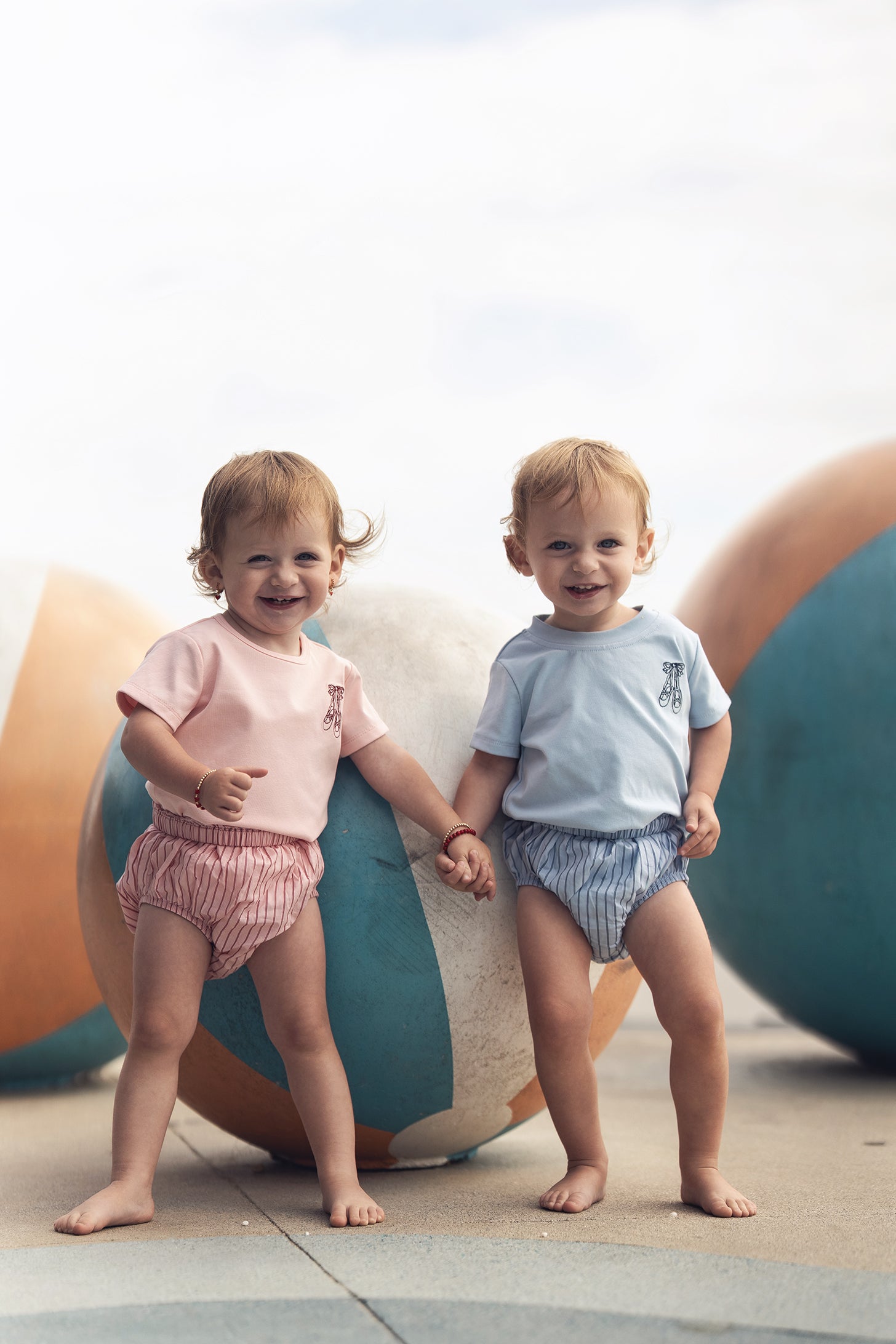 Two toddlers in pink and blue striped cotton bloomers, holding hands outdoors beside large colorful balls.