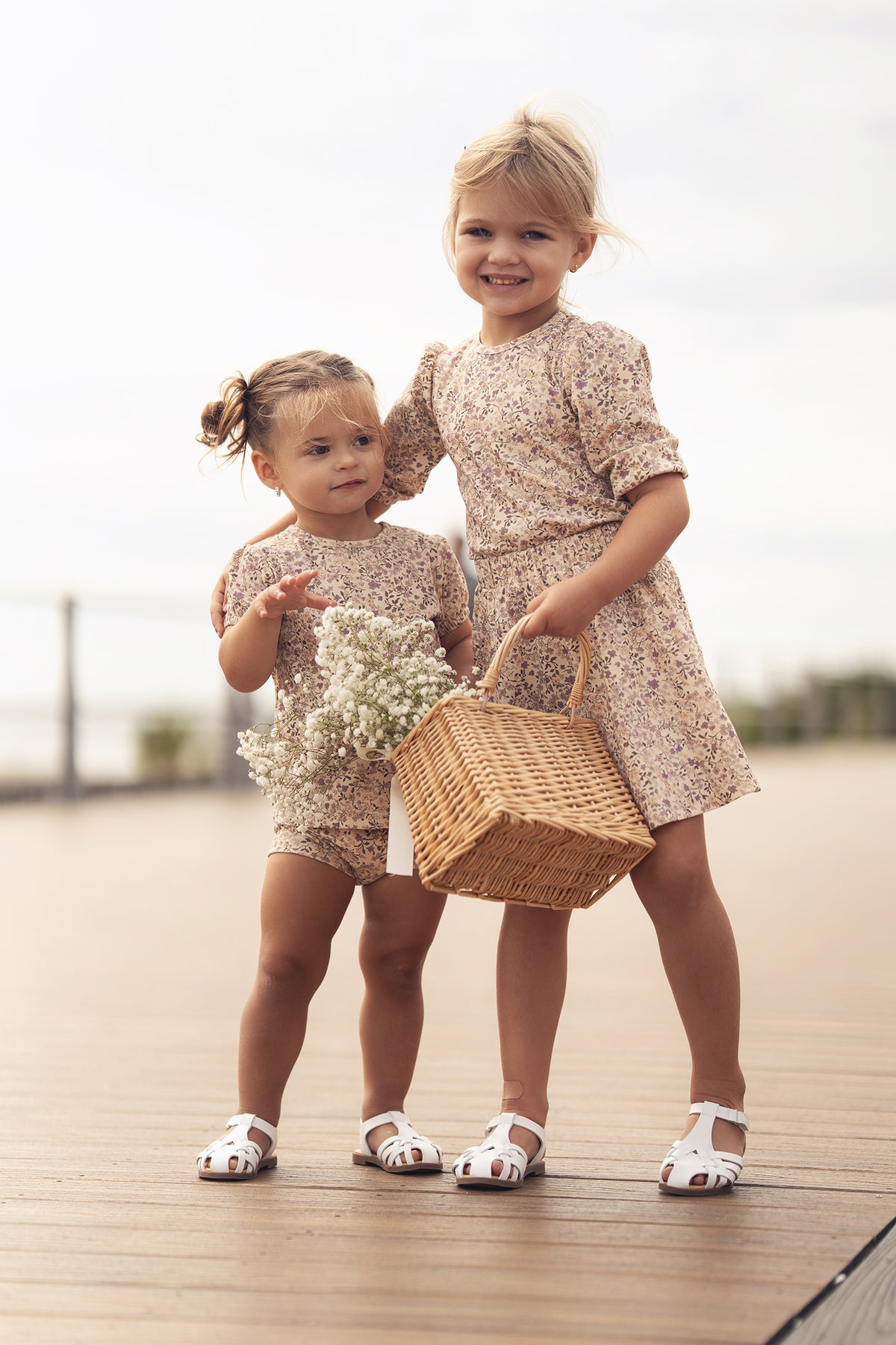 Two girls in purple flower skirt outfits on a boardwalk; one carries a wicker basket with flowers.