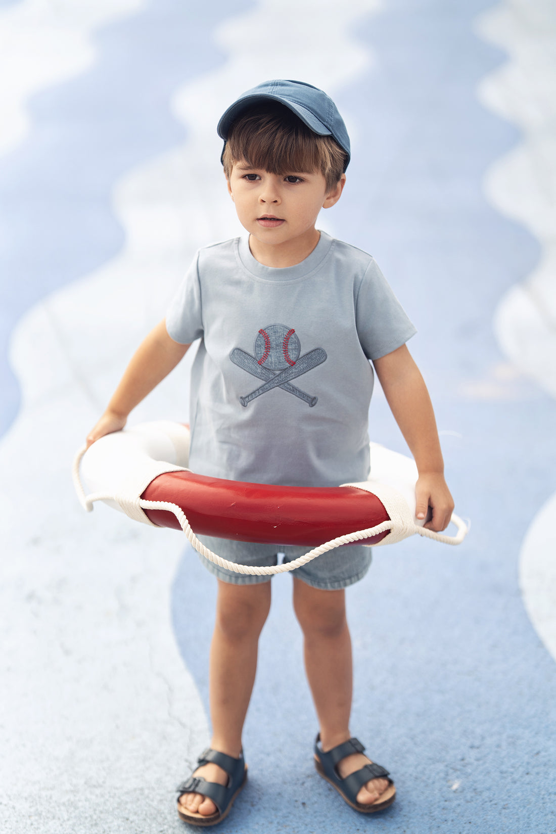 Child wearing light blue baseball T-shirt with crossed bats and ball graphic, blue cap.